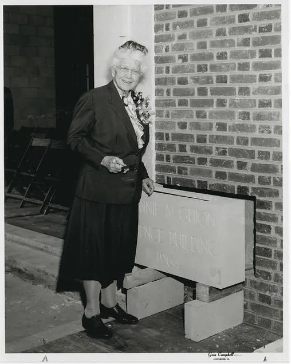 Older woman holding flowers, standing at a brick construction site, with a commemorative plaque that reads "John M. Gion S...
