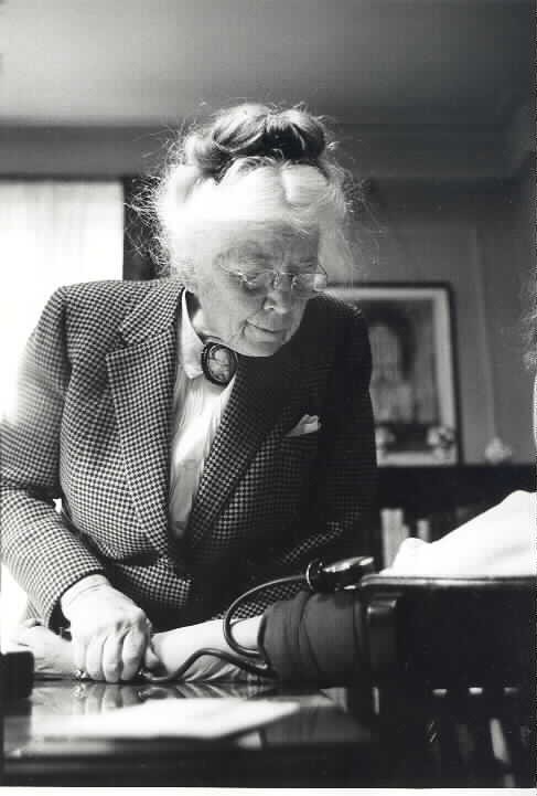 An elderly woman in a checkered coat uses a blood pressure cuff, possibly an early device used for this purpose, shown in ...